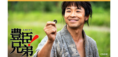 Actor Taiga Nakano holding a coin in period clothing for the NHK Taiga drama Toyotomi Kyodai against a grassy field.